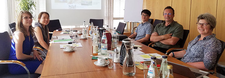 Fünf Mitarbeiter sitzen um einen langen Konferenztisch in einem gut beleuchteten Raum und blicken in Richtung eines Präsentationsbildschirms, der landwirtschaftliche Projekte in Baden-Württemberg zeigt.